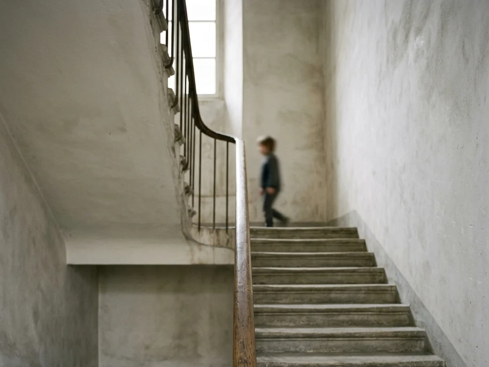 A quiet plaster stairwell with a worn wooden handrail and a figure walking up past a bright window.